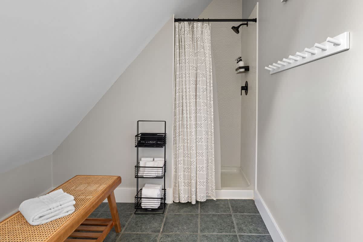 A compact bathroom with a sloped ceiling features a walk-in shower with a patterned curtain, a wooden bench, and a black metal storage rack, set against light gray walls and dark tile flooring.