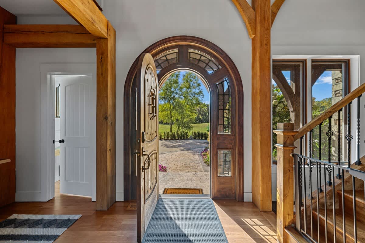 Grand entryway featuring an arched wooden door with intricate hardware, opening to a lush green outdoor landscape. Exposed timber beams frame the space, with a white door visible to an adjacent room.