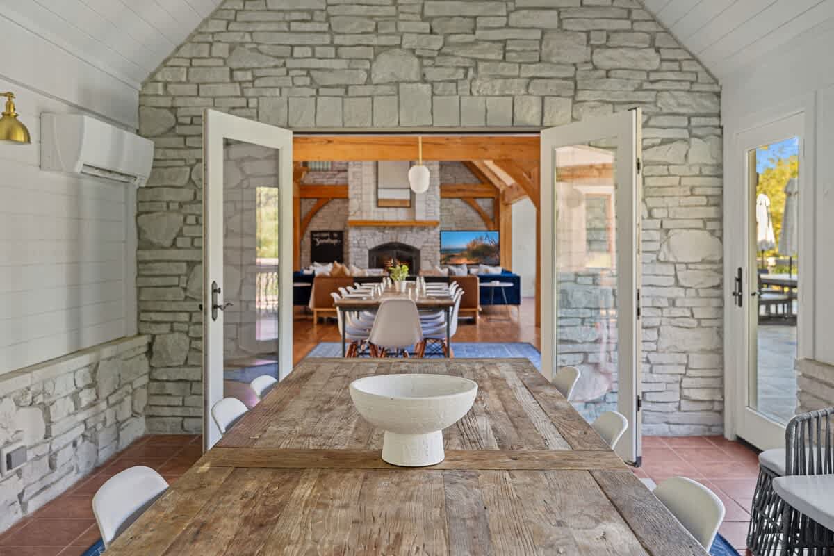 A rustic dining room with a long wooden table and white chairs, featuring stone walls and large glass doors that open to a living area with a stone fireplace and timber beams.