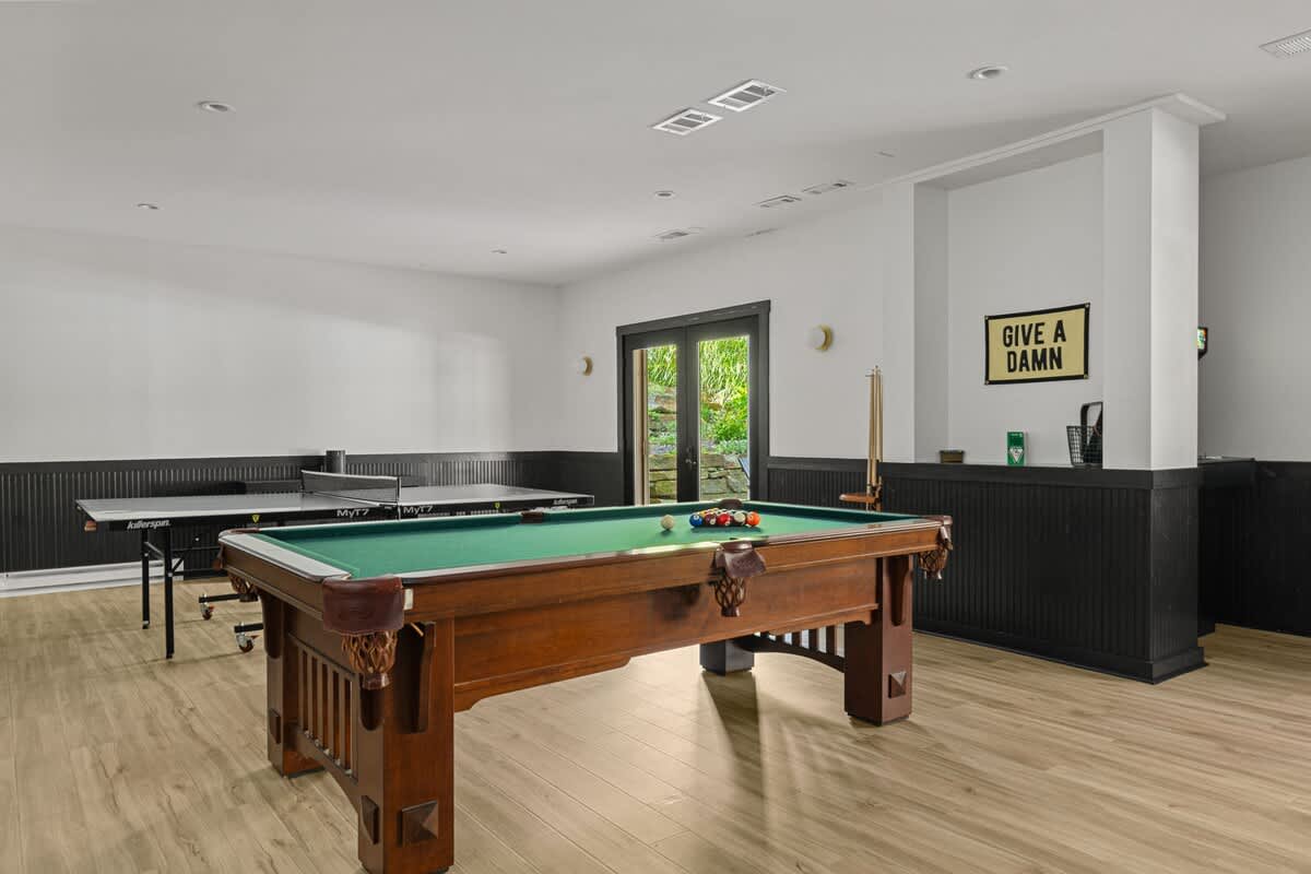 Expansive game room with light wood-look flooring and a black wainscoting accent wall, featuring a classic wooden pool table and a black ping pong table. A 'GIVE A DAMN' sign adds a playful touch.