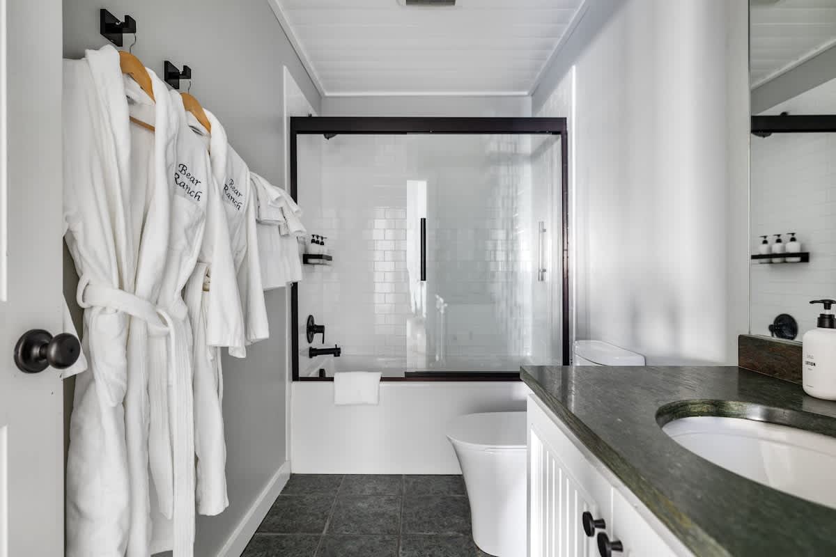 Modern bathroom with dark gray floor tiles, white subway tile shower, and a dark green countertop vanity. White bathrobes hang on the wall, adding a touch of luxury and comfort.