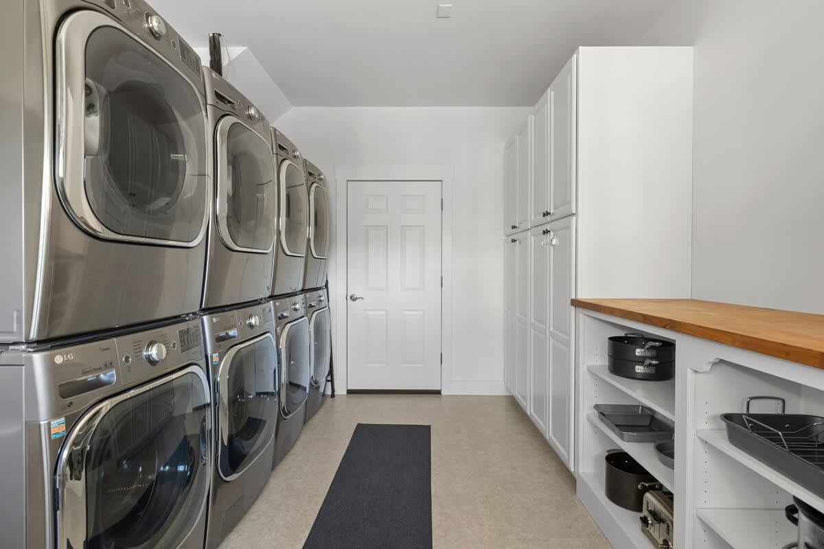 Spacious and well-lit laundry room equipped with multiple stacked silver washer and dryer units. White storage cabinets line one wall, complemented by a wooden countertop, and a dark rug on the floor.