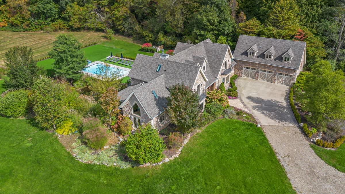 An elevated aerial view captures the large stone house with its complex roofline and dormers, nestled within a lush, treed landscape. The outdoor pool area and a long gravel driveway are also visible.
