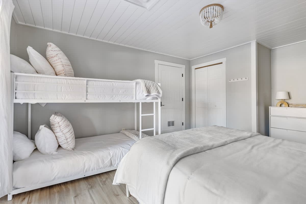 Cozy bedroom with light wood-look flooring and light gray walls, featuring a white bunk bed and a queen bed, both with white bedding. A small dresser with a lamp is visible in the corner.