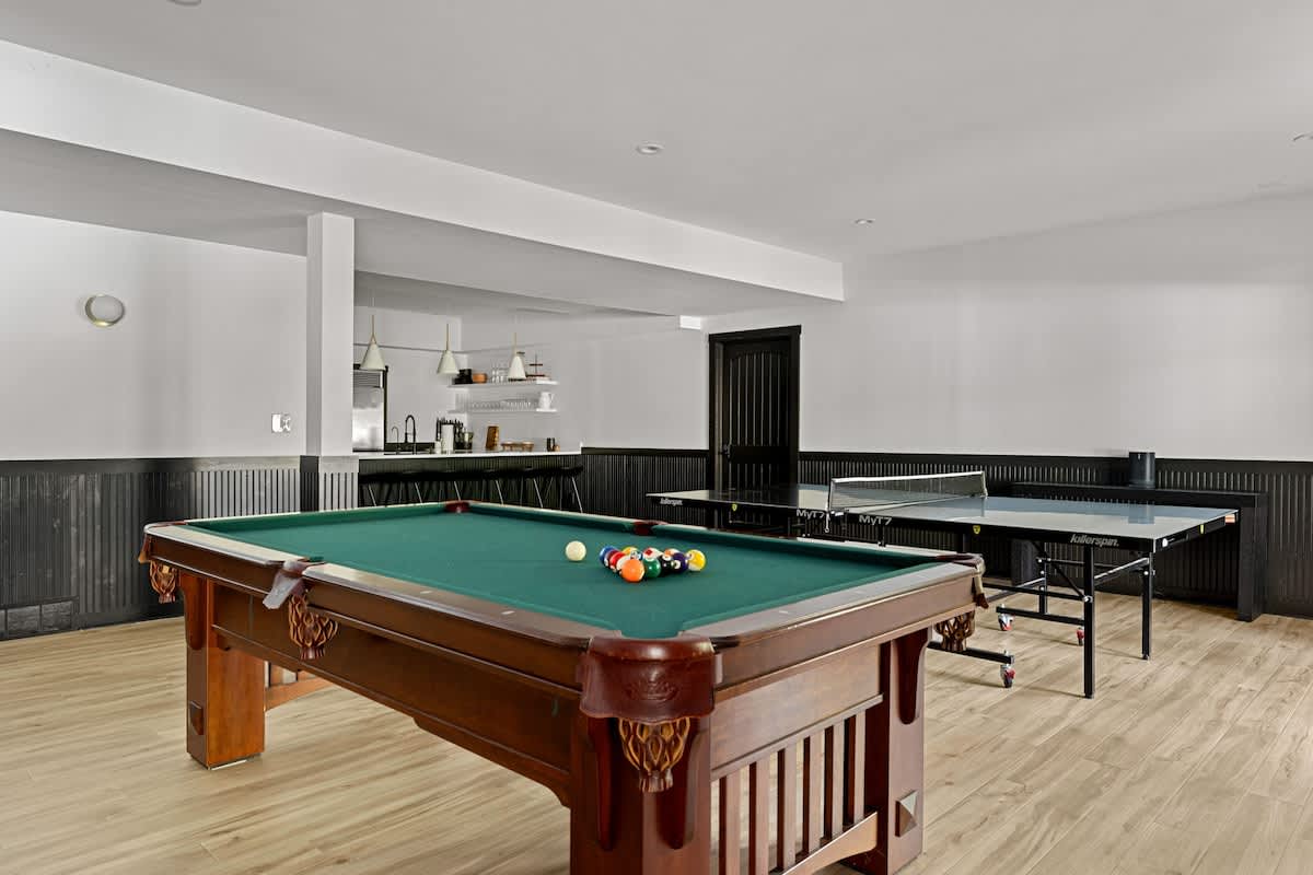 A spacious game room offers entertainment with a classic pool table and a ping pong table, set on light wood-look flooring, with a modern bar area featuring open shelving and pendant lights in the background.