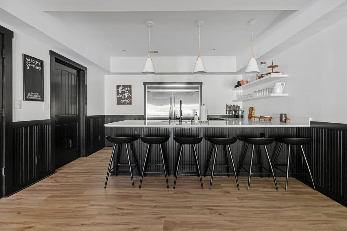 Modern wet bar or kitchenette area with a long white countertop, black bar stools, and a large sink with a commercial-style faucet. The space features black wainscoting and light wood-look flooring.