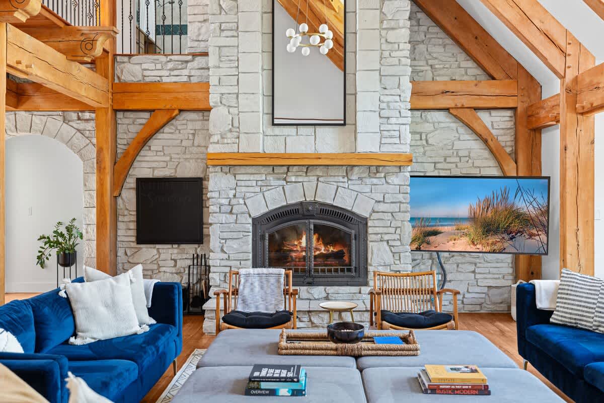 Close-up view of the living room's impressive stone fireplace with a roaring fire, flanked by two stylish wooden chairs. A large wall-mounted television is visible to the right, complementing the exposed timber beams.
