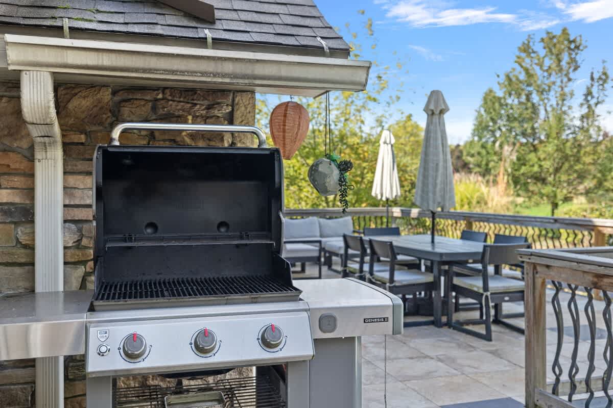 Stainless steel BBQ grill positioned on a tiled outdoor deck, ready for use. The background shows the outdoor dining and lounge areas, surrounded by green foliage and trees under a bright sky.