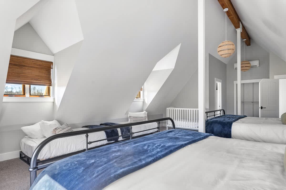 A spacious attic bedroom with sloped white ceilings and light gray walls features multiple black metal-frame beds with blue throws, a crib, and a woven pendant light.