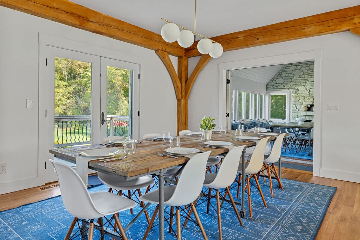 Dining room with a long wooden table, a mix of white and grey chairs, and a striking blue patterned rug. French doors lead to an outdoor area, and an arched doorway connects to another stone-walled room.