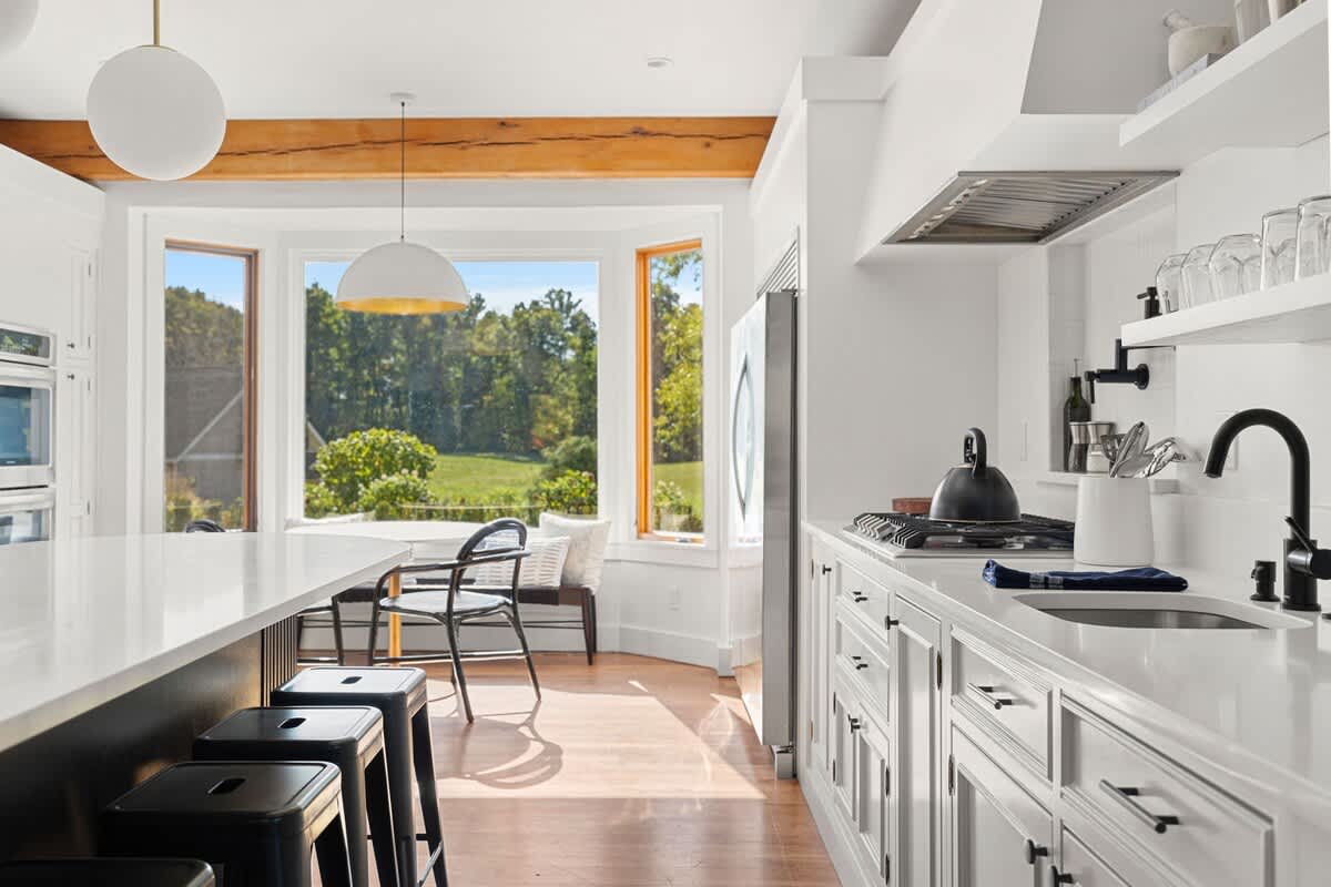 A modern kitchen view showing white cabinets, a gas range with a stainless steel hood, and a large black kitchen island with white countertops, leading to a bright bay window with a small table.