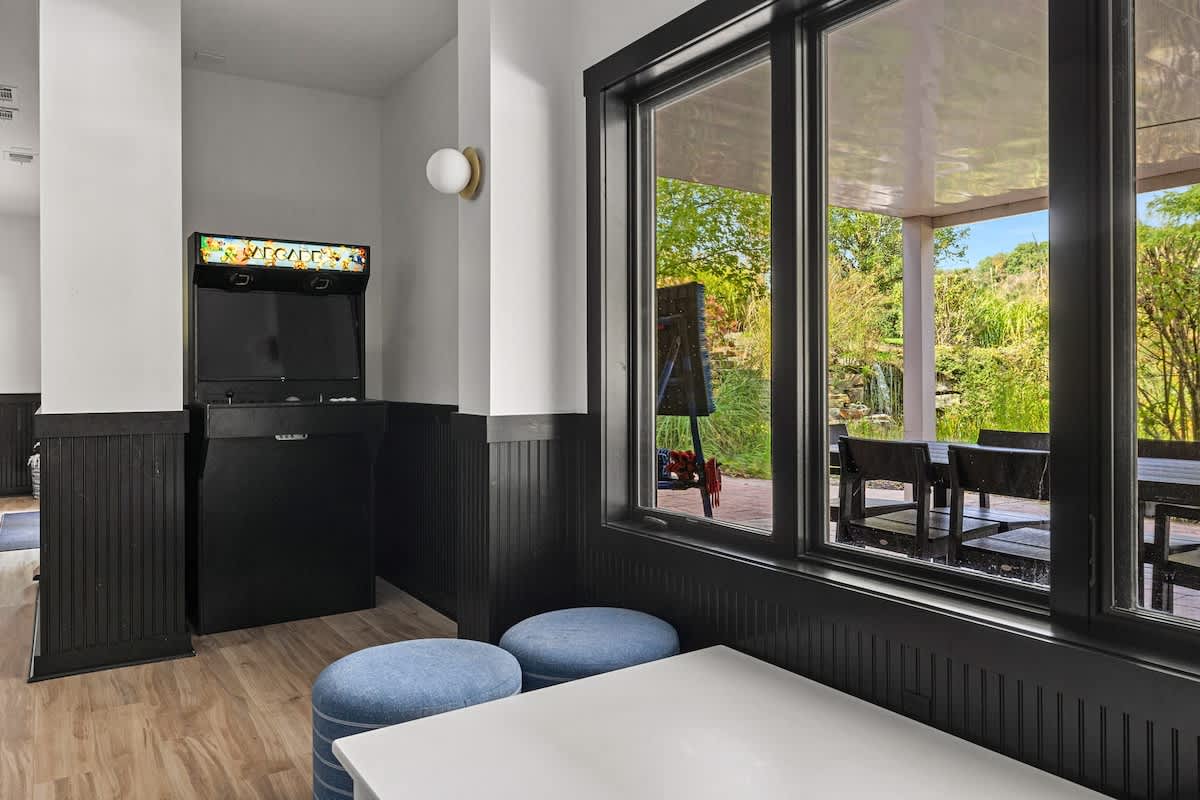 Section of a game room with an arcade machine and large windows overlooking an outdoor patio. Light wood-look flooring and black wainscoting continue the room's aesthetic, with blue ottomans providing seating.