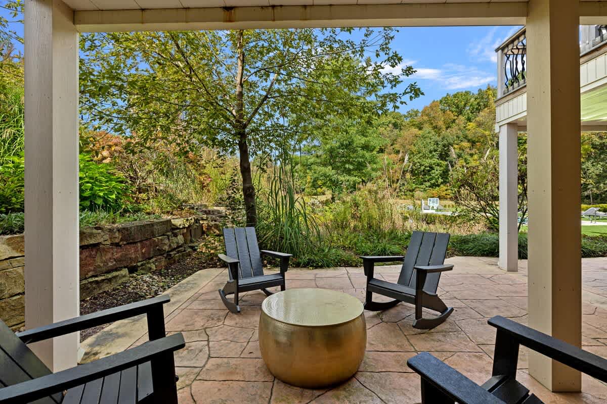 A covered outdoor patio features a stamped concrete floor, two dark rocking chairs, and a round gold coffee table, overlooking a lush green landscape with trees and a stone retaining wall.