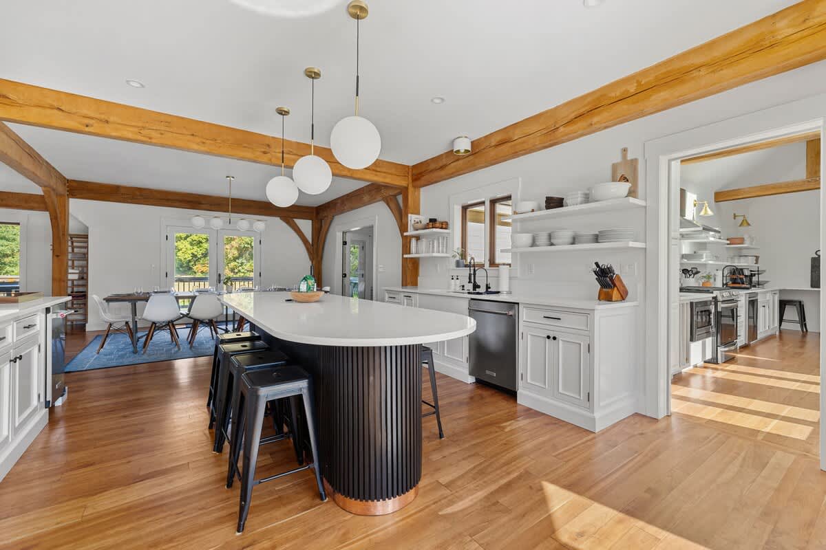 An expansive open-concept kitchen with white cabinetry, a large black island, and exposed wooden beams, flowing into a dining area with a blue rug and a table, all on hardwood floors.