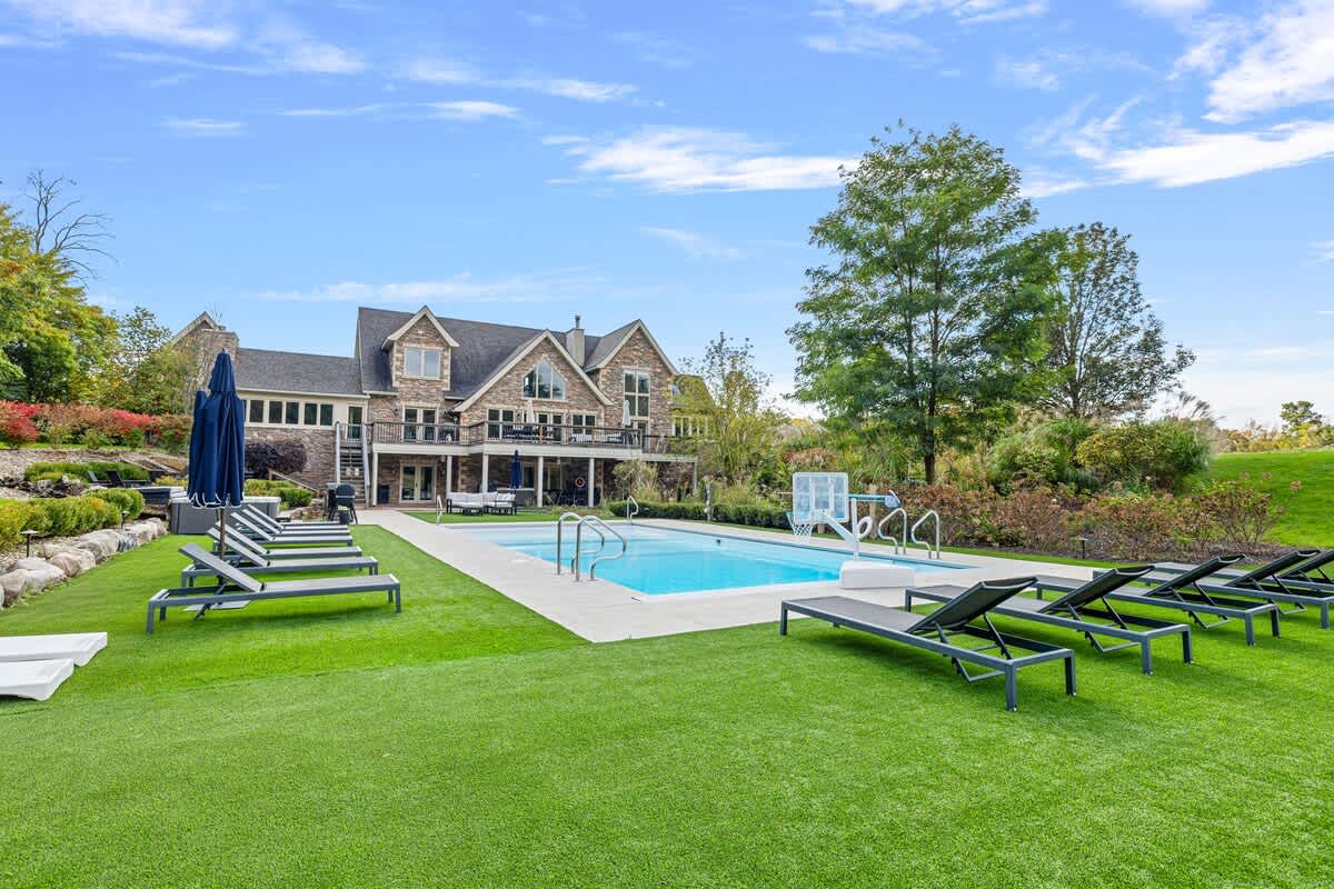 The back of a large stone and siding house with multiple gables overlooks a rectangular outdoor pool, surrounded by artificial turf and numerous lounge chairs. Mature trees and natural landscaping frame the expansive property.