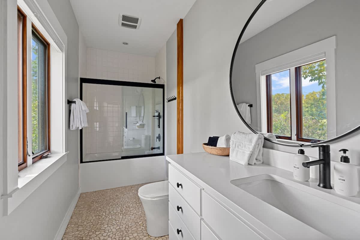 Modern bathroom with a white vanity, black faucet, and a large round mirror. It features a shower/tub combo with a black-framed glass enclosure and pebble-like floor tiles, with a window providing natural light.