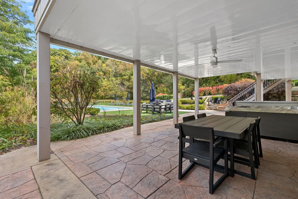 A spacious covered patio with a stamped concrete floor hosts a dark outdoor dining set and a built-in hot tub, offering views of a distant swimming pool and lush greenery.