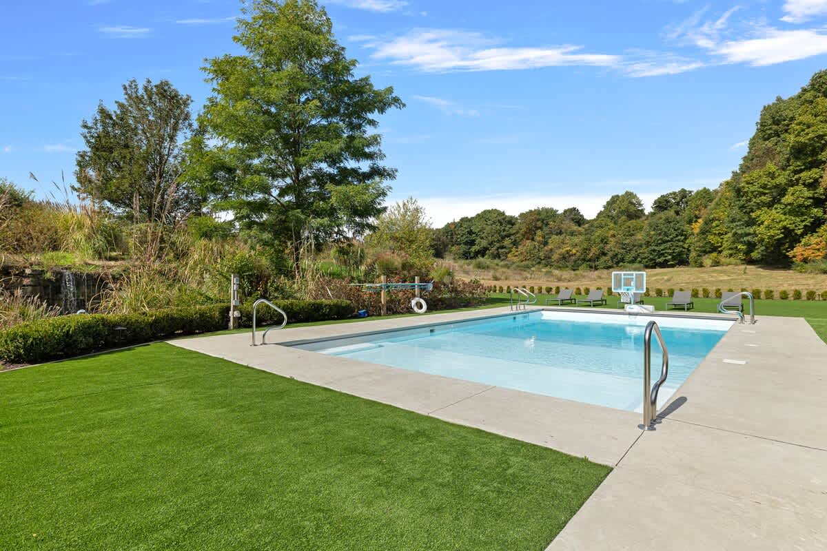 An outdoor swimming pool with concrete decking is surrounded by lush artificial turf, backed by a natural landscape of trees and a small waterfall feature on a grassy hill.