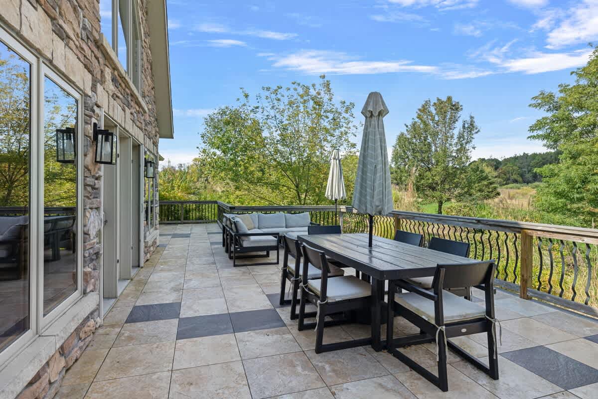 Expansive outdoor deck with patterned tile, featuring both a dining area with an umbrella and a comfortable lounge seating area. The deck is adjacent to a stone-clad exterior and offers views of lush green trees and a distant pool.