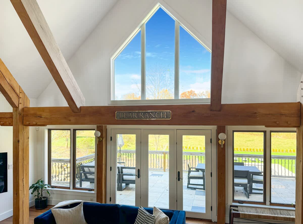 Bright living space featuring a high vaulted ceiling and expansive windows that offer panoramic views of the outdoors. A 'BEAR RANCH' sign is mounted above the French doors leading to an outdoor deck.