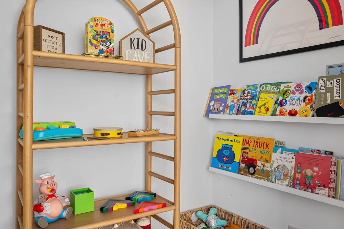 Dedicated kid's play corner with a wooden arch-shaped bookshelf filled with toys and a wall-mounted shelf displaying children's books. A colorful rainbow art print hangs above, creating a cheerful space.