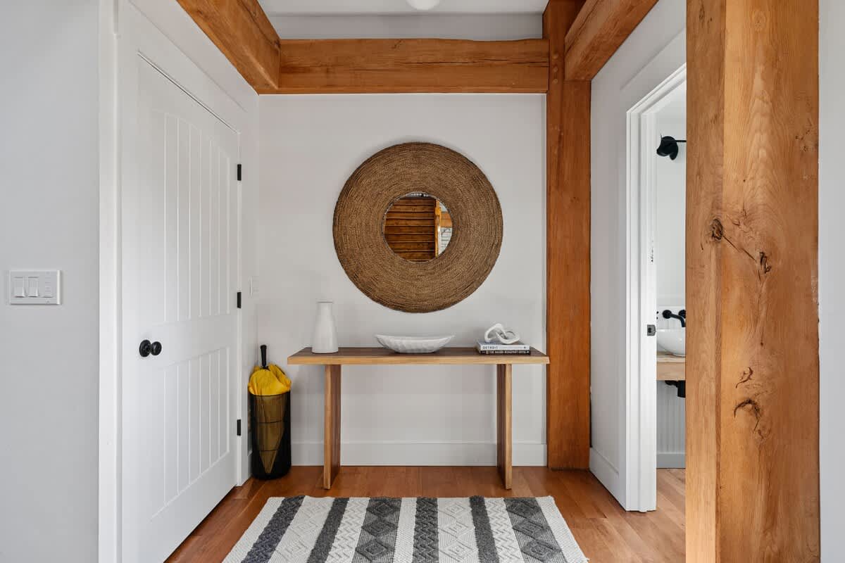 A welcoming foyer with light hardwood floors and exposed wooden beams, featuring a rustic wooden console table adorned with decor and a large circular woven mirror above it.