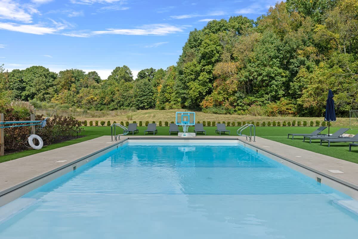 A clear, rectangular outdoor swimming pool with a light blue interior and concrete coping is shown, featuring a pool basketball hoop and surrounded by artificial turf.