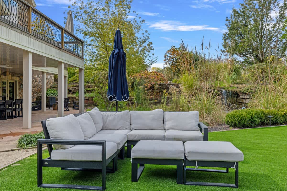 A modern outdoor sectional sofa with light gray cushions and an ottoman sits on a manicured lawn, next to a large blue umbrella, with a view of the house's lower deck and lush garden.