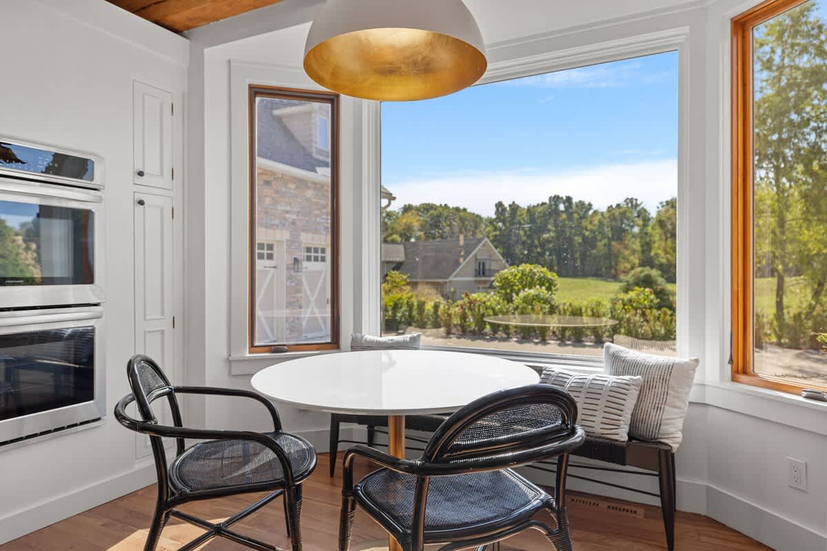 A cozy breakfast nook within the kitchen, featuring a round white table, two black chairs, and a built-in bench with pillows, all set against large windows offering views of the exterior.