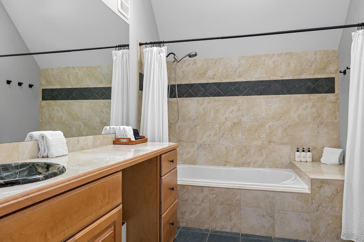 Spacious bathroom with a large built-in bathtub, light-colored tiled walls with a dark accent stripe, and a double vanity with a marble countertop. A shower curtain encloses the tub area, and white towels are neatly stacked.