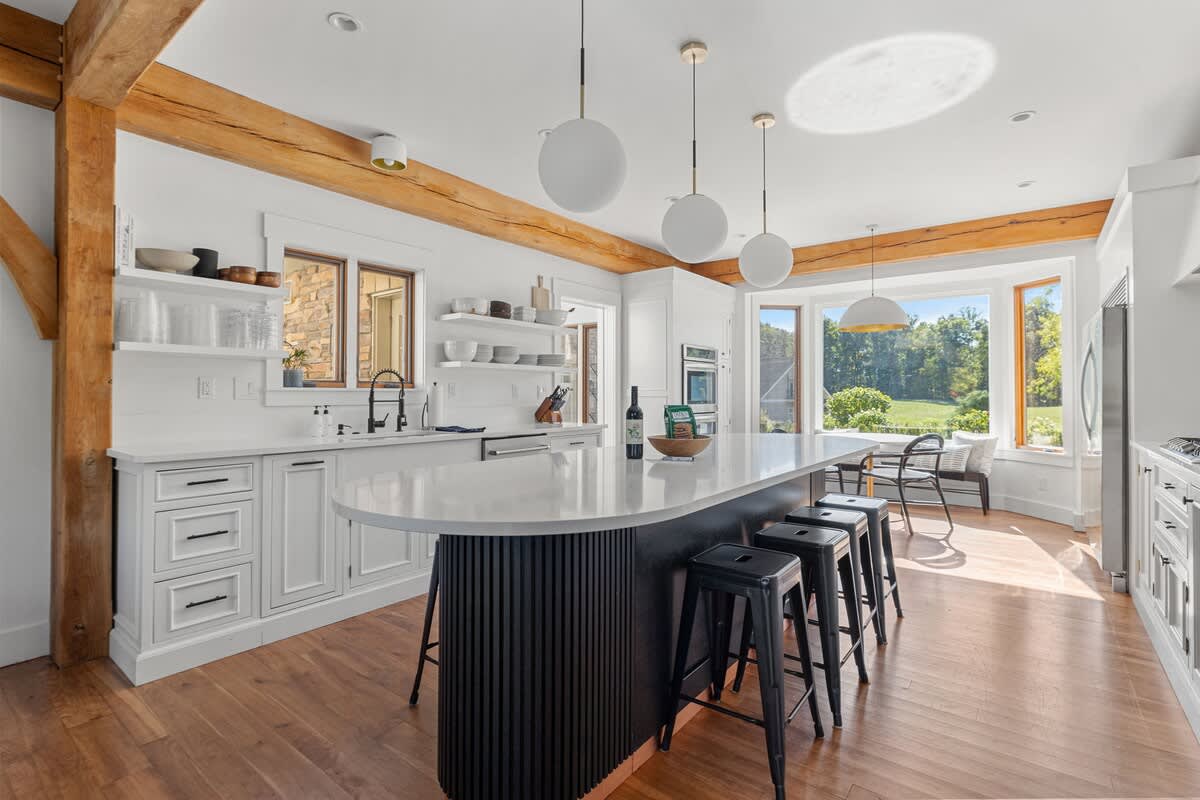 A spacious, bright kitchen with white cabinets, a large black island with white stools, and exposed wooden beams, featuring stainless steel appliances and a bay window with a small seating area.