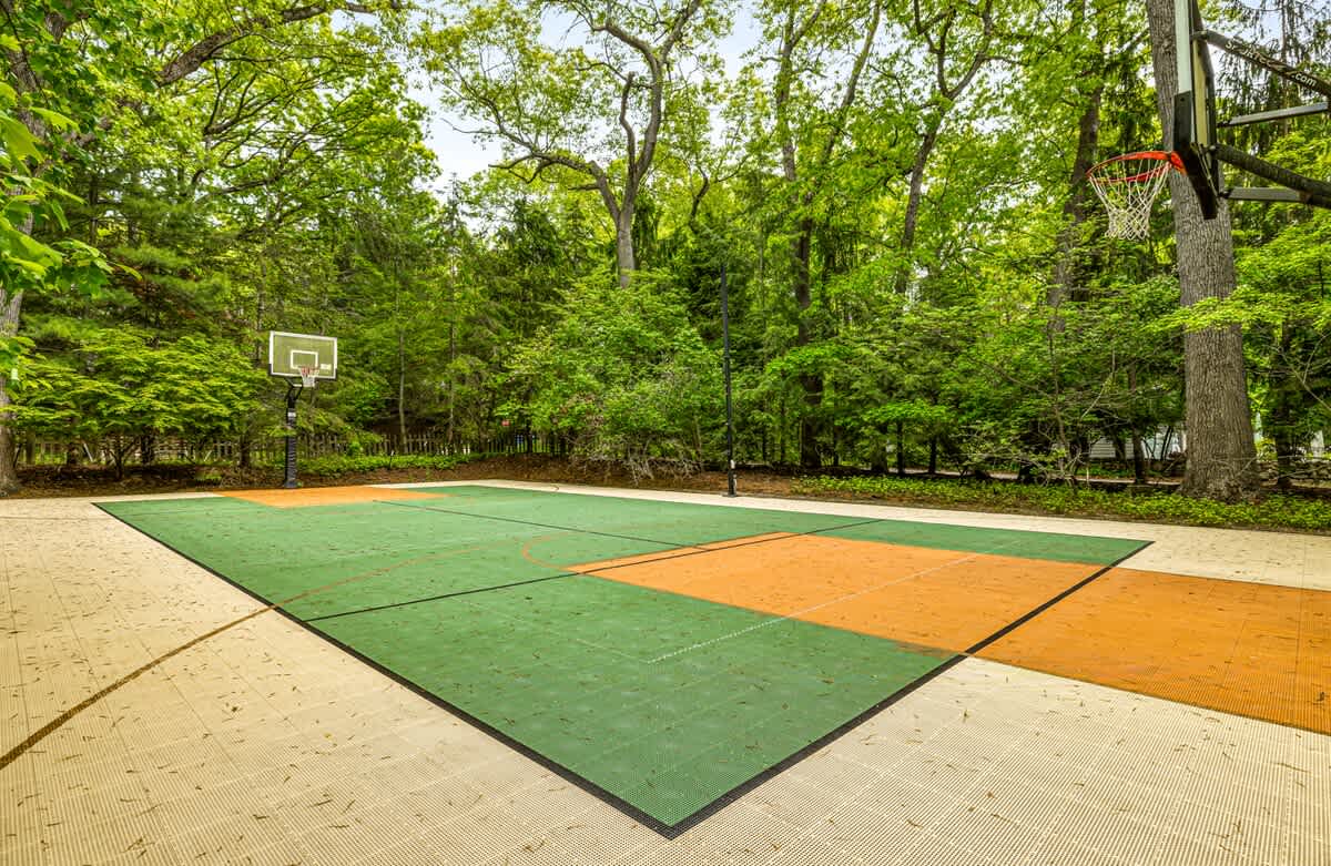 An outdoor sports court with green and orange interlocking tiles, featuring two basketball hoops, set amidst a lush green forest with tall trees. The court is surrounded by lighter-colored paving.