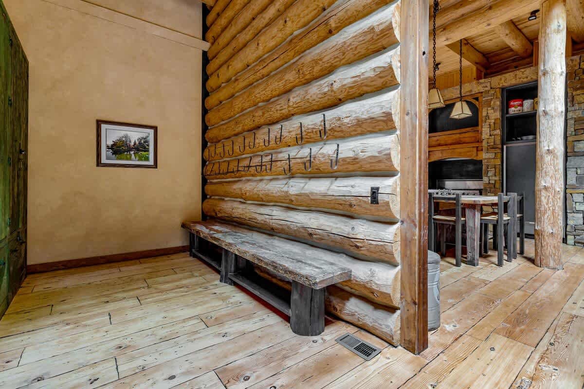 A functional mudroom area in a log cabin, featuring a long wooden bench, numerous coat hooks on the exposed log wall, and light wood flooring.
