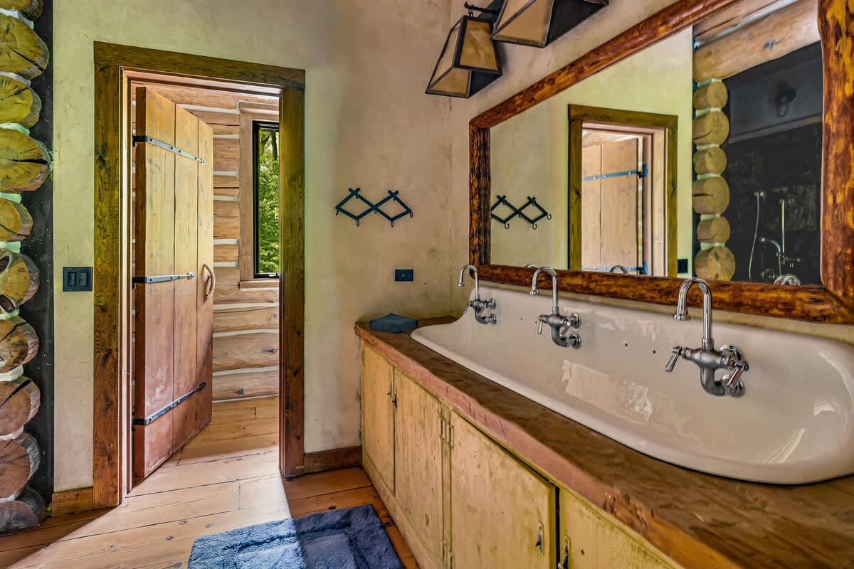 Rustic bathroom featuring a long white trough sink with multiple faucets, set into a wooden countertop and vanity, with a large log-framed mirror above and a wooden door leading to a log-walled room.