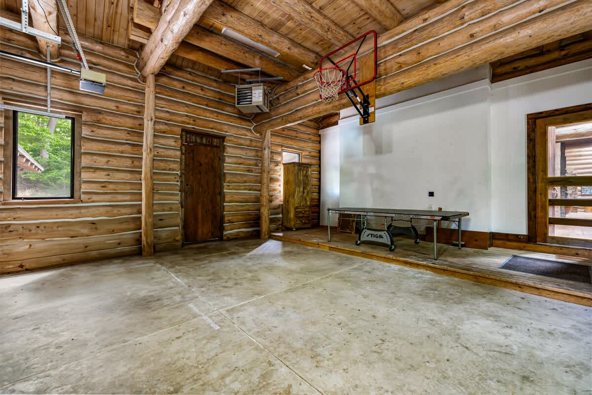 The interior of a spacious log cabin garage, featuring a concrete floor, exposed log walls, a basketball hoop, and a ping pong table, suggesting a dual-purpose recreation space.