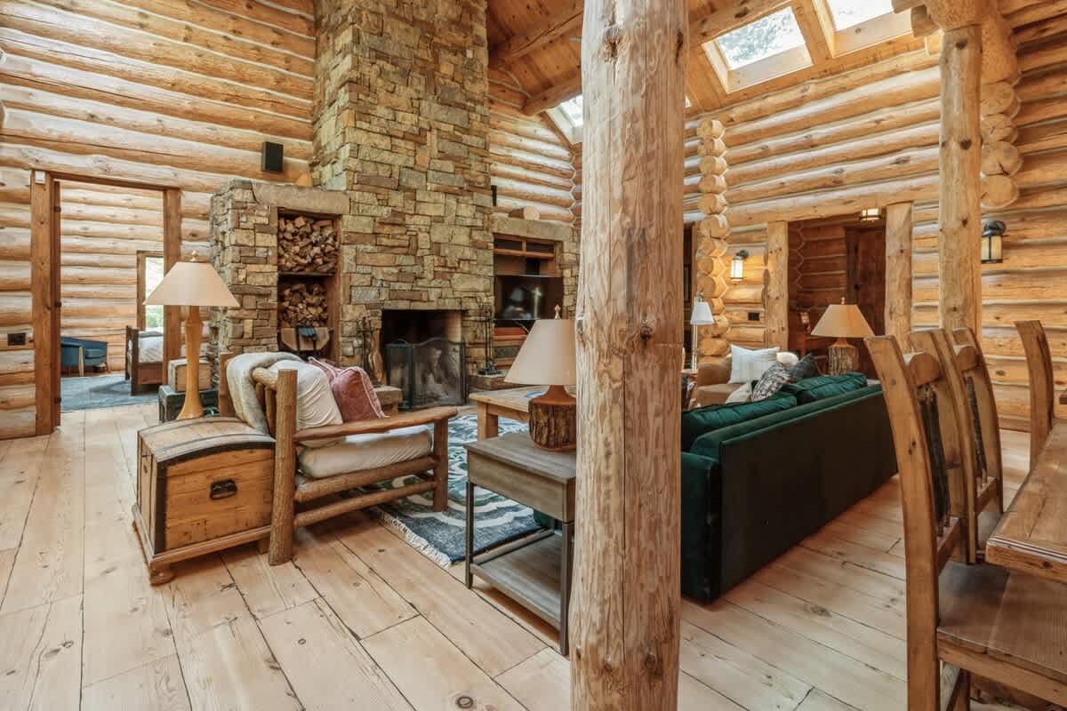 A cozy log cabin living room featuring a prominent stone fireplace, exposed log walls and ceiling, light wood floors, and comfortable seating including a green sofa and a rustic armchair.