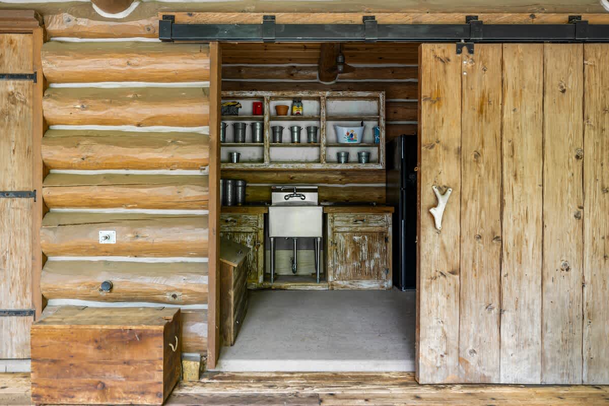 A rustic outdoor utility area or mudroom with a large metal sink, open shelving stocked with various items, and a black refrigerator, all within a log cabin structure with a sliding barn door.