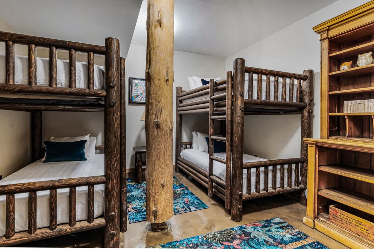 Spacious bunk room featuring two sets of rustic log bunk beds with white bedding and blue pillows, separated by a large wooden support beam, with colorful patterned rugs on a concrete floor.