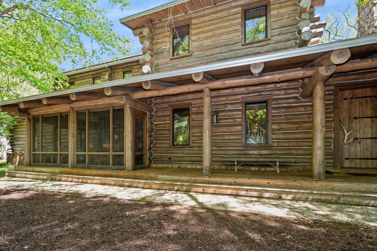 The exterior of a charming log cabin featuring a long screened porch with wooden framing and a wide front porch with wooden railings, surrounded by mature trees and natural landscaping.