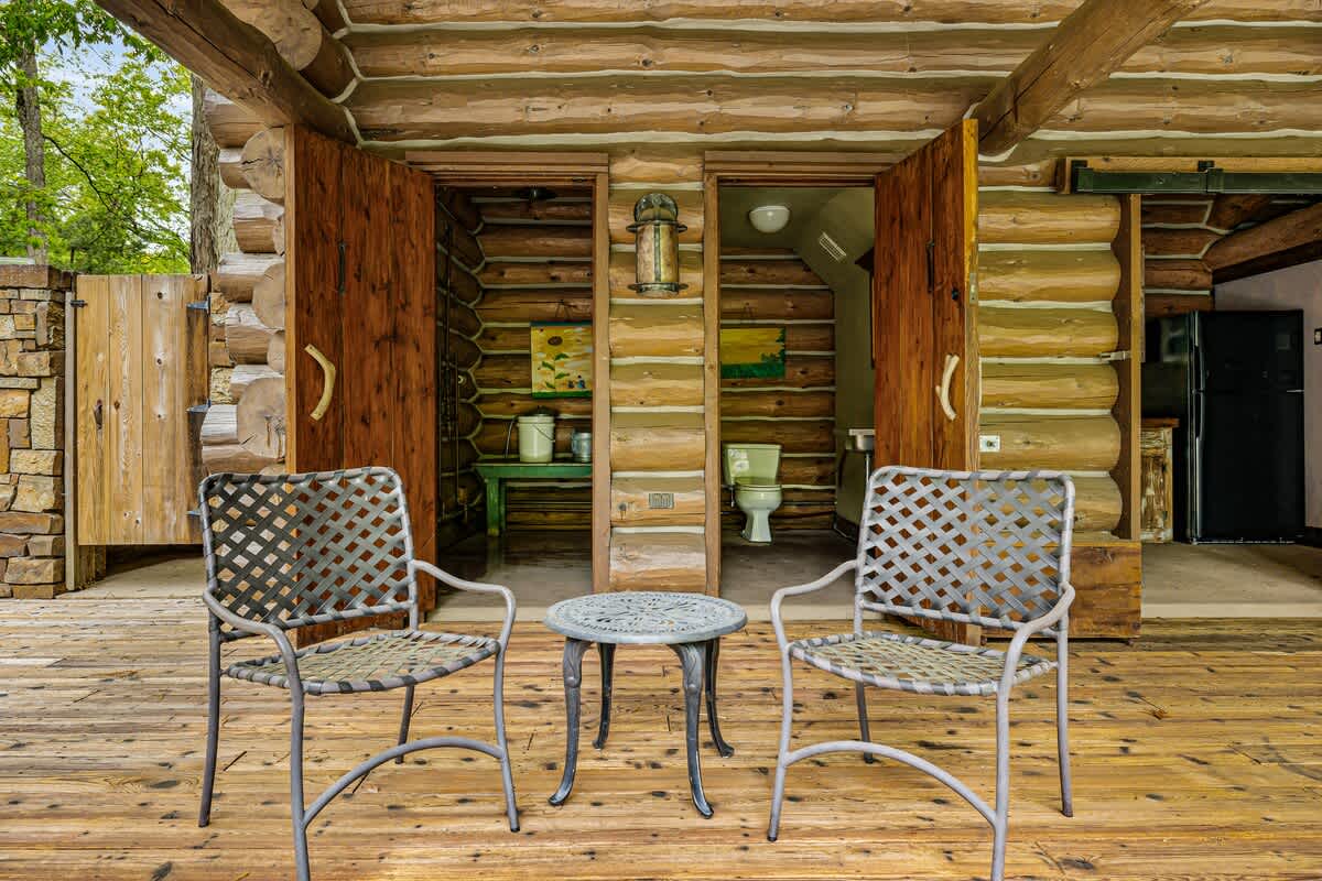 A rustic wooden deck features two metal chairs and a small round table, positioned outside two wooden doors leading into a log-walled interior. One door reveals a bathroom with a toilet, suggesting a convenient pool house setup.