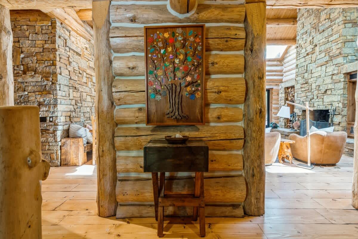 A rustic log cabin interior hallway with exposed log walls, light wood floors, and a decorative wooden tree art piece hanging above a small wooden console table.