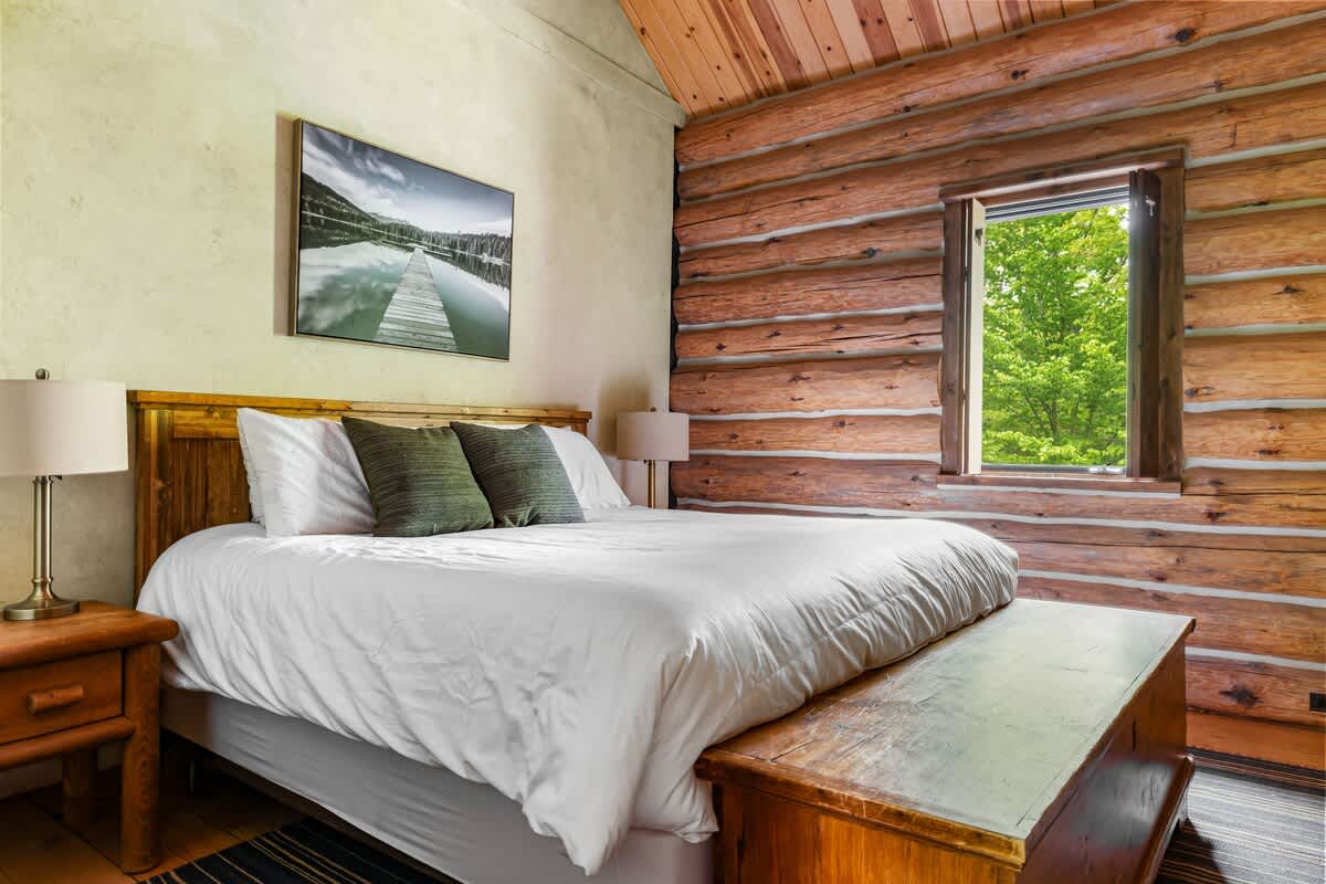 Cozy bedroom featuring a large bed with white bedding, a wooden headboard, and two dark green pillows, set against a light green textured wall and rustic log walls with a window overlooking lush greenery.