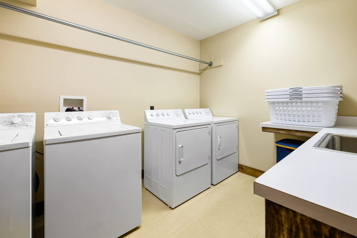 A bright laundry room features two sets of white top-loading washers and front-loading dryers, with a long metal hanging rod above. A white countertop with a sink and stacked laundry baskets provides a functional workspace.
