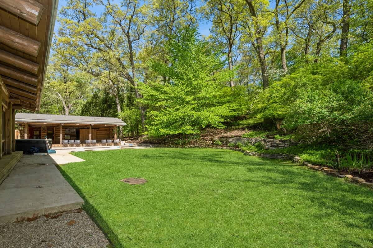 A well-maintained green lawn in a backyard setting, bordered by a concrete patio and lush trees, with a log cabin structure visible in the distance.