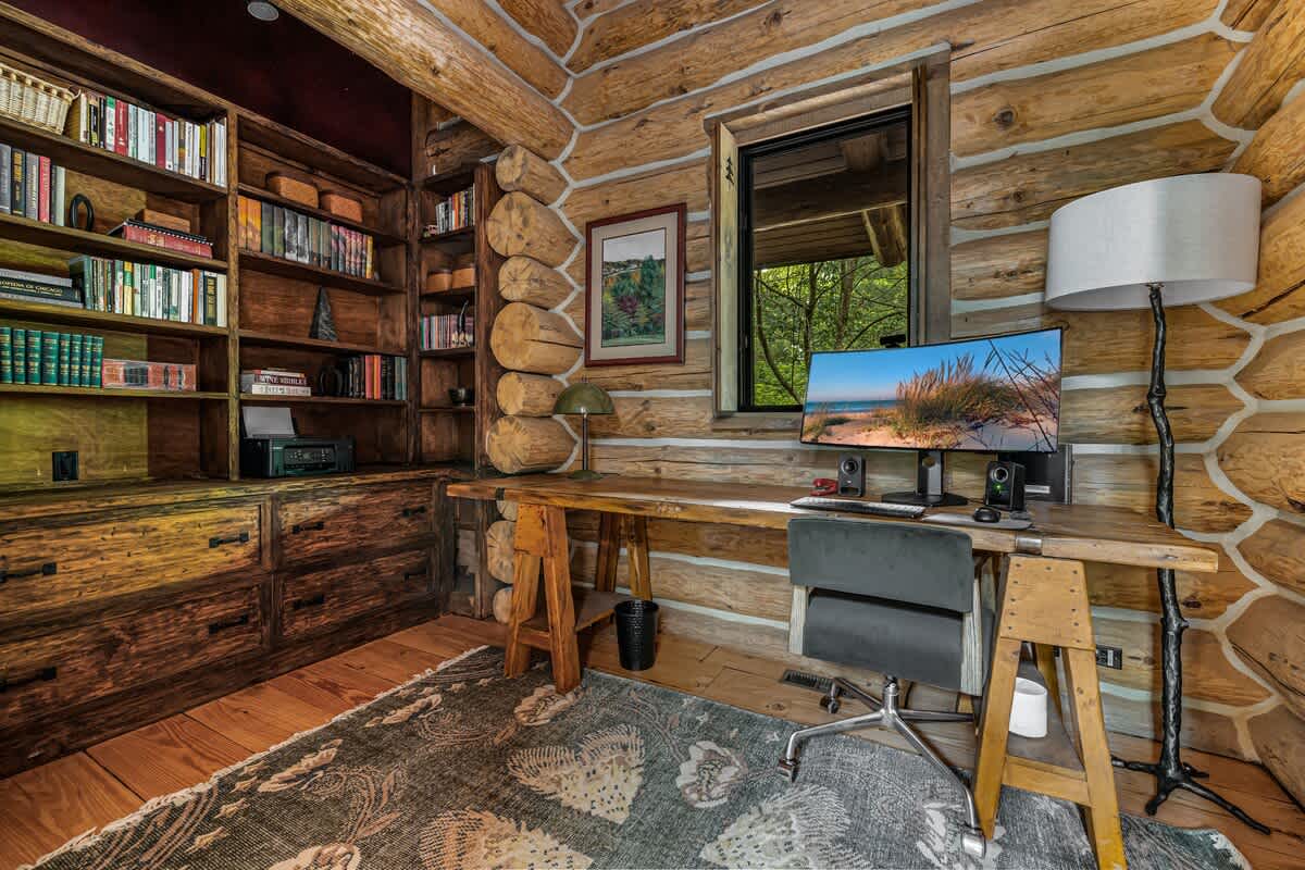 Cozy log cabin office with a large wooden desk, ergonomic chair, and a computer monitor, complemented by built-in wooden bookshelves filled with books and a patterned area rug.