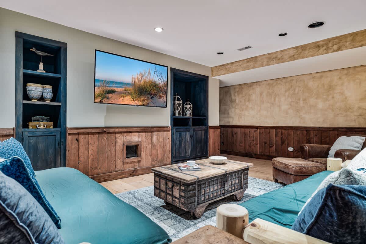 A cozy media room features a large wall-mounted TV above a rustic wood-paneled wall, flanked by dark blue built-in shelves. Two teal sofas face a distressed wooden coffee table, creating a comfortable lounging area.