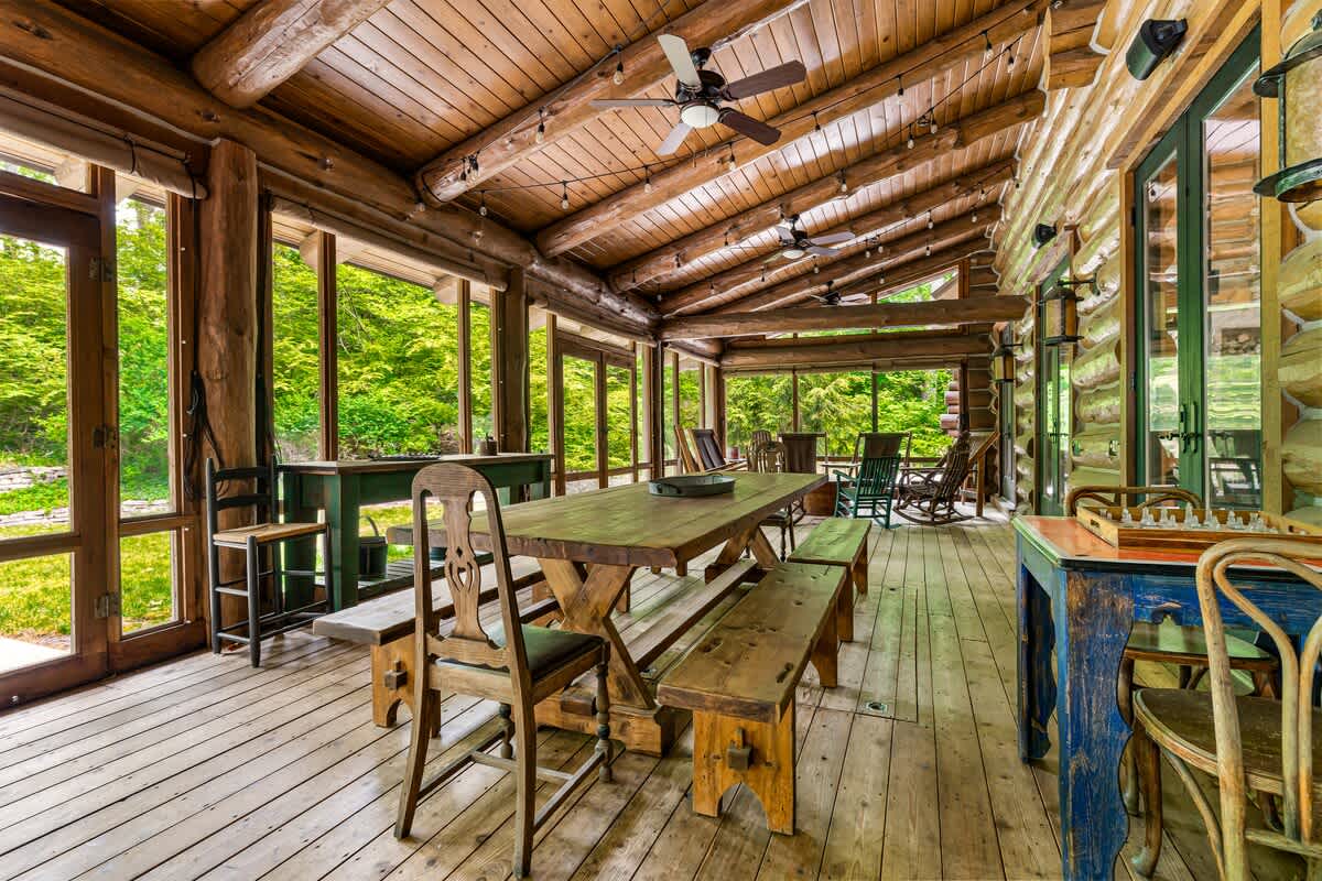 A spacious screened-in porch or sunroom with a high vaulted wooden ceiling, multiple ceiling fans, and a large rustic wooden dining table with benches, surrounded by lush greenery visible through the screens.