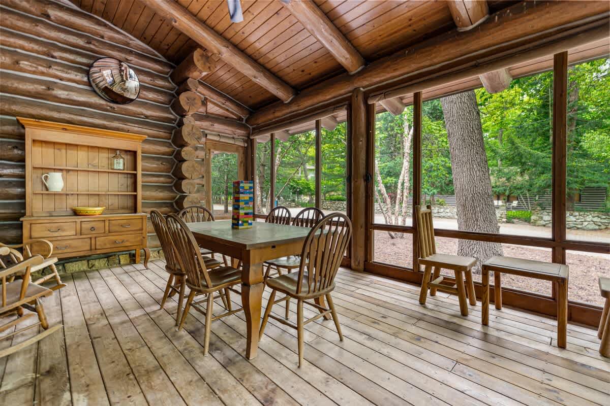 Rustic screened-in porch featuring log cabin walls, a wooden plank floor, and a large dining table with six wooden chairs, all surrounded by expansive windows offering views of lush greenery.