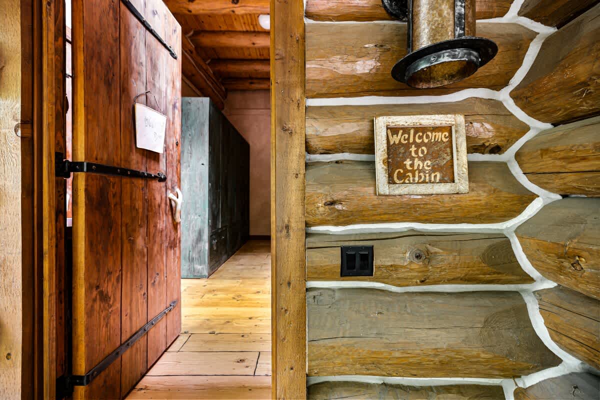 A rustic interior entryway featuring exposed log walls with white chinking, a sturdy wooden door with black hardware, and a 'Welcome to the Cabin' sign, leading into a hallway with wooden floors.