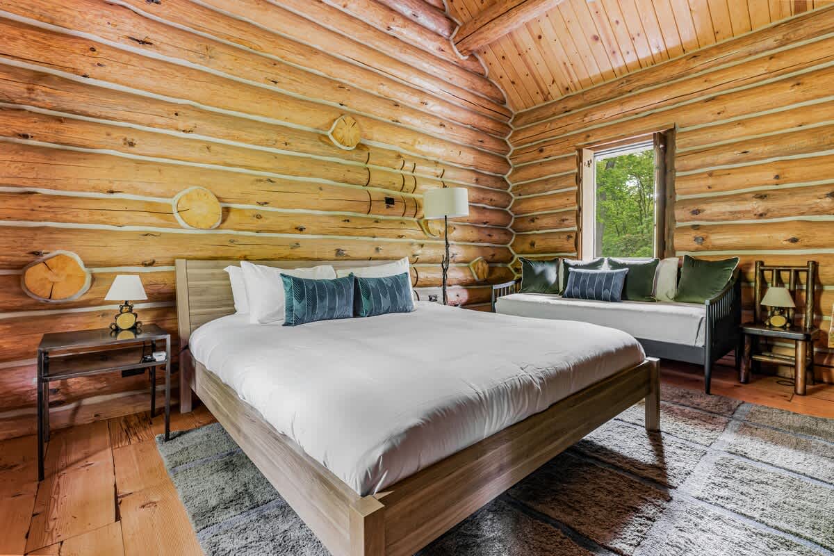 Cozy bedroom with exposed log walls and ceiling, featuring a large bed with teal pillows, a daybed, and a window overlooking green trees.